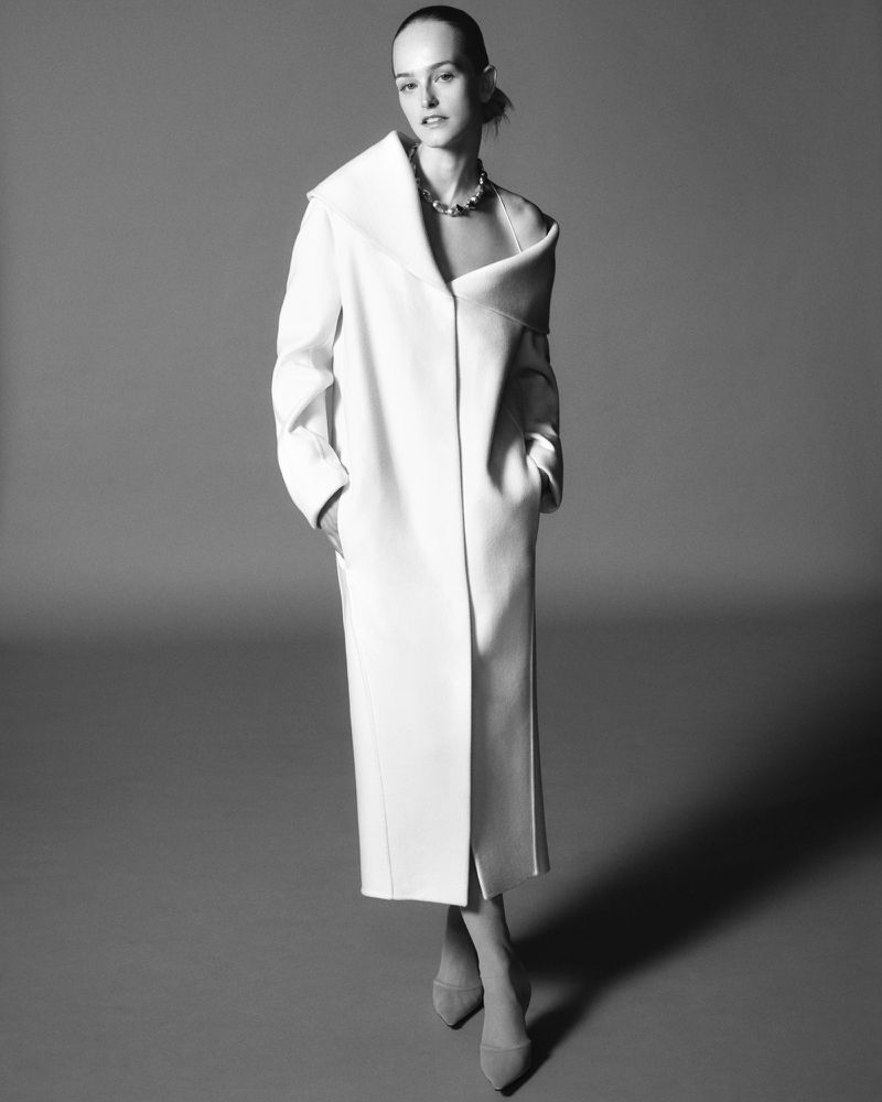 Model wearing a white structured coat and statement necklace against a grey gradient studio background