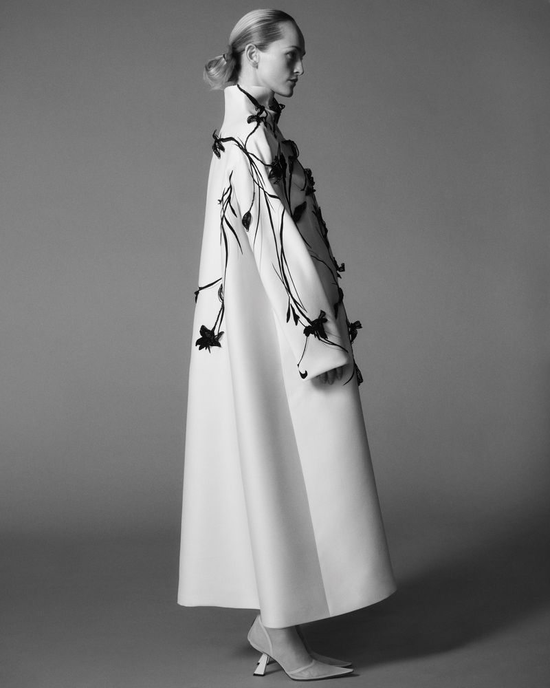 Monochrome fashion portrait: a slender adult female with blonde hair in an elegant white coat adorned with black floral details and high heels.