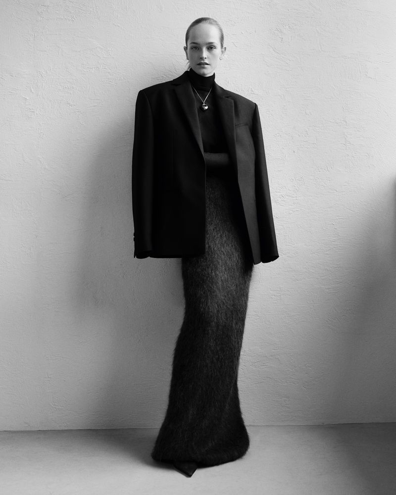 A slender woman in a black turtleneck, oversized blazer, and shaggy skirt poses against a textured neutral wall in a full-length portrait.
