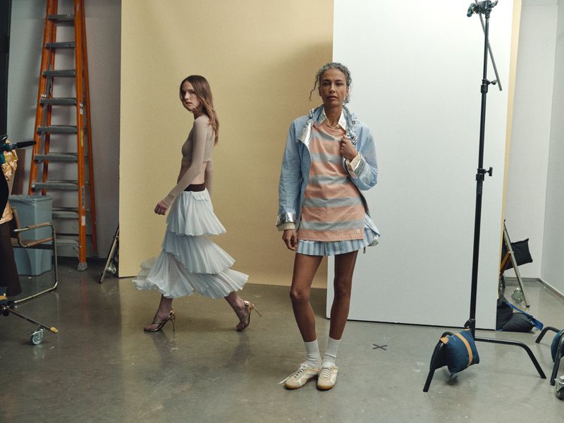 Two women in a studio setting. One walks in a tiered skirt, while the other poses in a striped top and jacket in soft studio light.