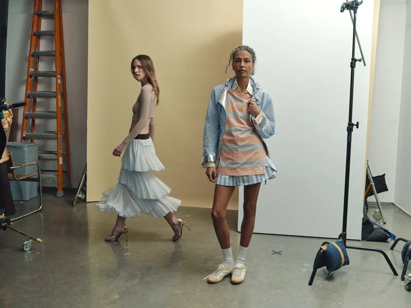 Two women in a studio setting. One walks in a tiered skirt, while the other poses in a striped top and jacket in soft studio light.