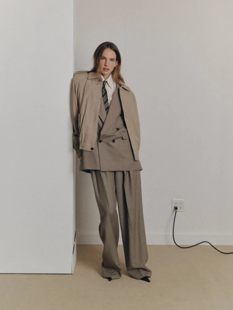 Fashionable woman in oversized suit and trench coat poses against a white wall, complemented by a striped tie and beige backdrop.