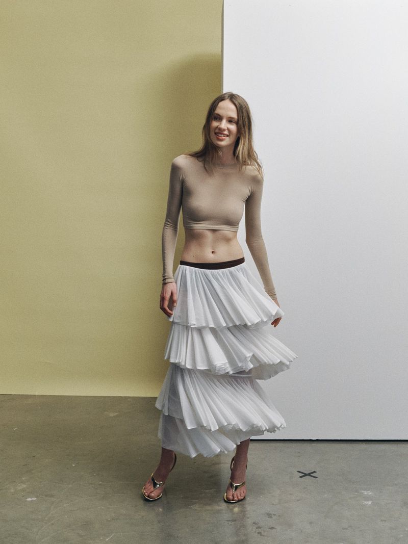 A slender woman with long brown hair poses in a studio setting, wearing a long-sleeved beige crop top and a tiered white skirt.