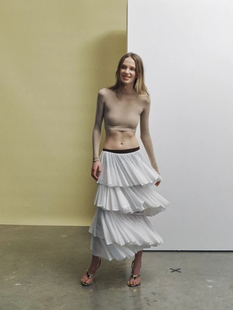 A slender woman with long brown hair poses in a studio setting, wearing a long-sleeved beige crop top and a tiered white skirt.