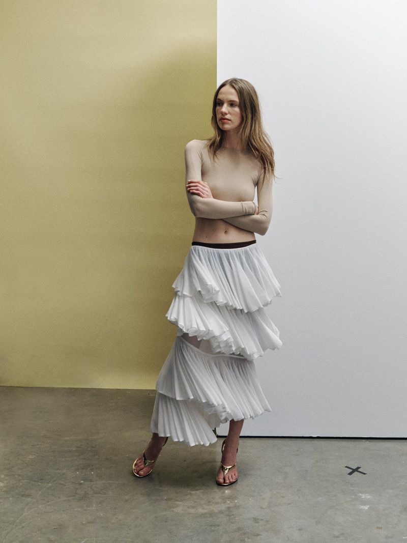 A woman poses indoors wearing a long-sleeved shirt, a tiered skirt, and heeled sandals, standing against a half yellow, half white wall.