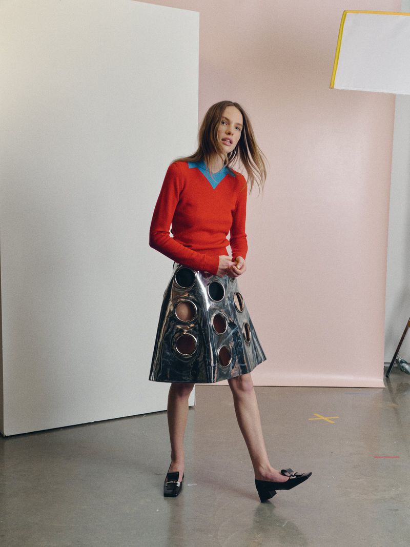 A woman in a red sweater and a metallic skirt with cutouts poses in a studio with a white and pink backdrop and studio lighting.