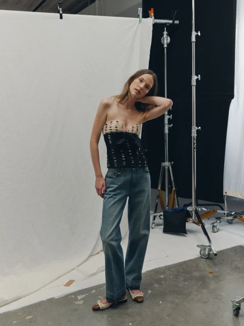 Full shot of a slender woman in a strapless top and jeans posing in a photo studio against a white backdrop with lighting equipment.
