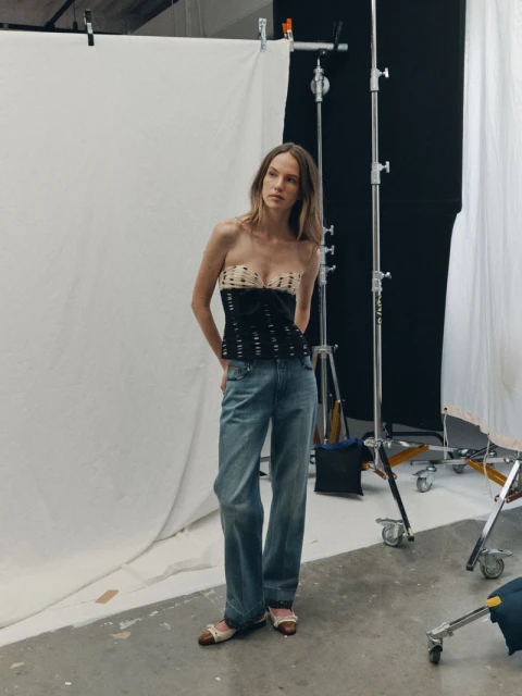 A fair-skinned young woman poses in a photo studio wearing a strapless top and baggy jeans, captured in a medium shot with soft lighting.