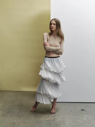 Model with long brown hair wearing beige long-sleeve top and white tiered pleated skirt against split olive and white background