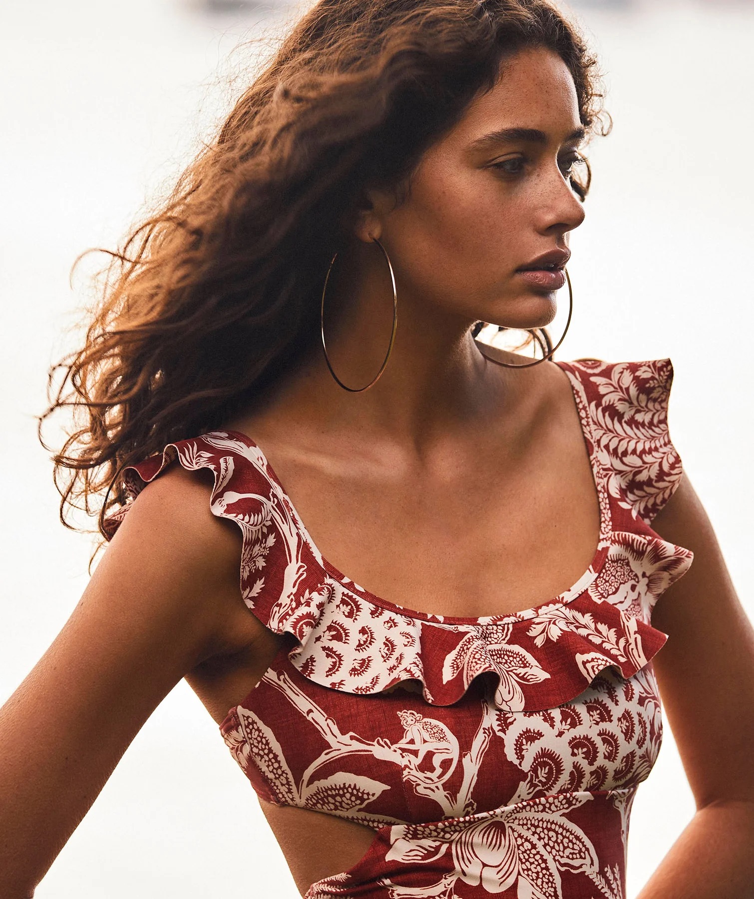 Adult woman wearing a patterned one-piece swimsuit with ruffle trim and large hoop earrings looking away from the camera.