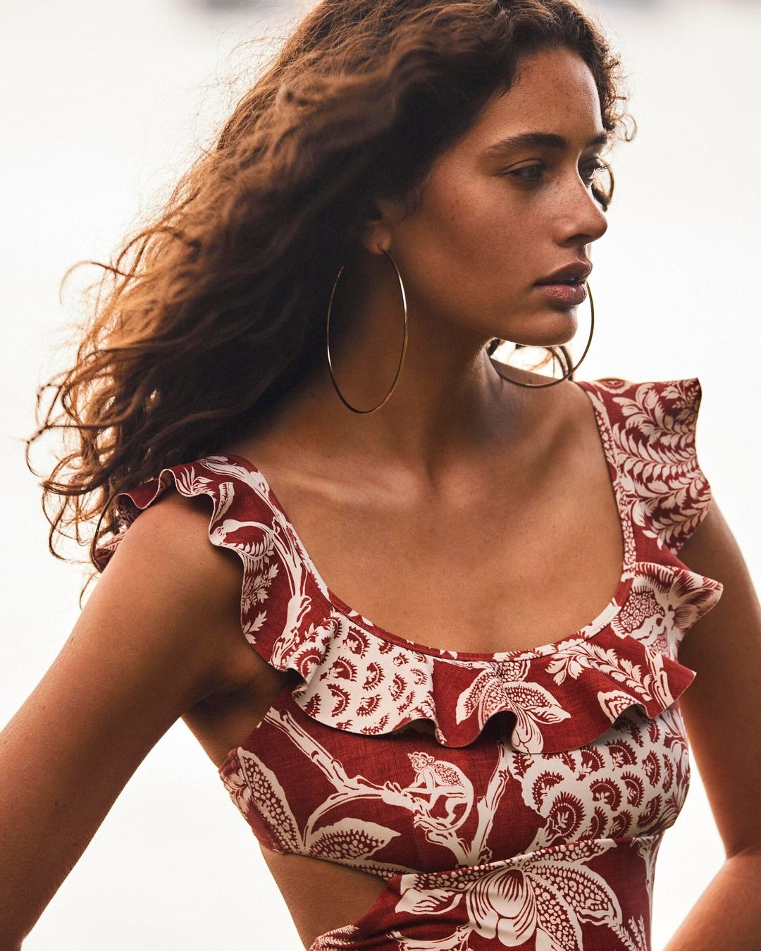 Close-up of a woman wearing a printed red and white patterned swimsuit and large gold hoop earrings against a bright outdoor background.