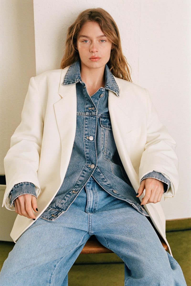 Model in denim jacket and oversized white blazer sitting against a neutral wall for Zara The Summer Edition