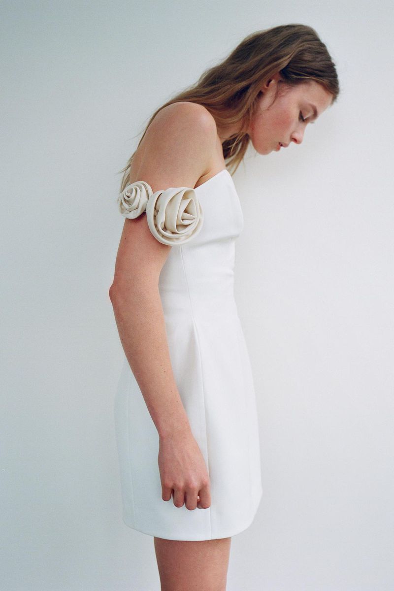 A young woman poses indoors wearing a white mini dress featuring floral arm embellishments against a neutral background.