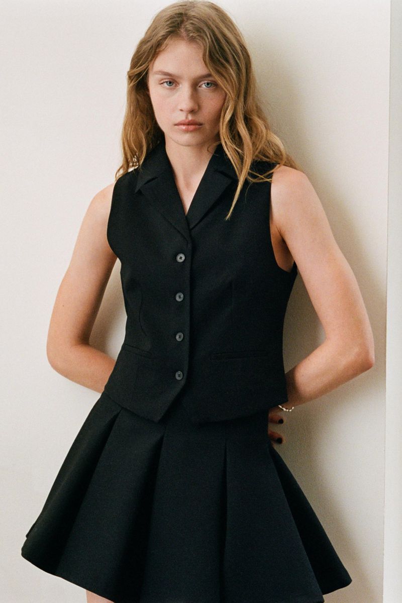 Model in a black buttoned vest and matching pleated skirt against a plain white studio wall for Zara.