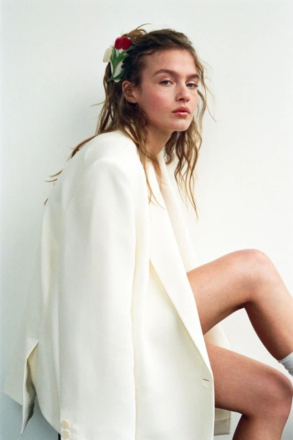 Portrait of a young woman in a white blazer with floral hair accessories, gazing at the camera with neutral expression against a white wall.