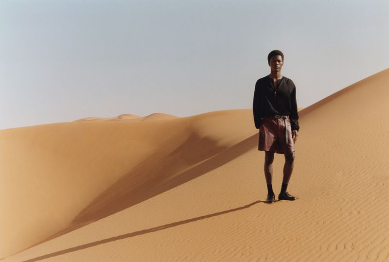 Adult male posing on a sand dune wearing a black long sleeve shirt and dark red shorts with a sand dune background under a clear sky.