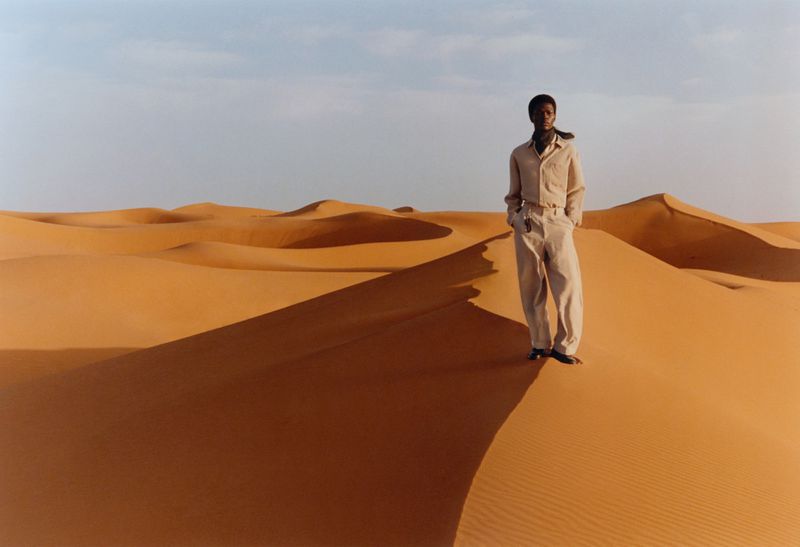 An adult male poses in a desert landscape, wearing a light-colored shirt and trousers, under a clear sky, standing on a sand dune.