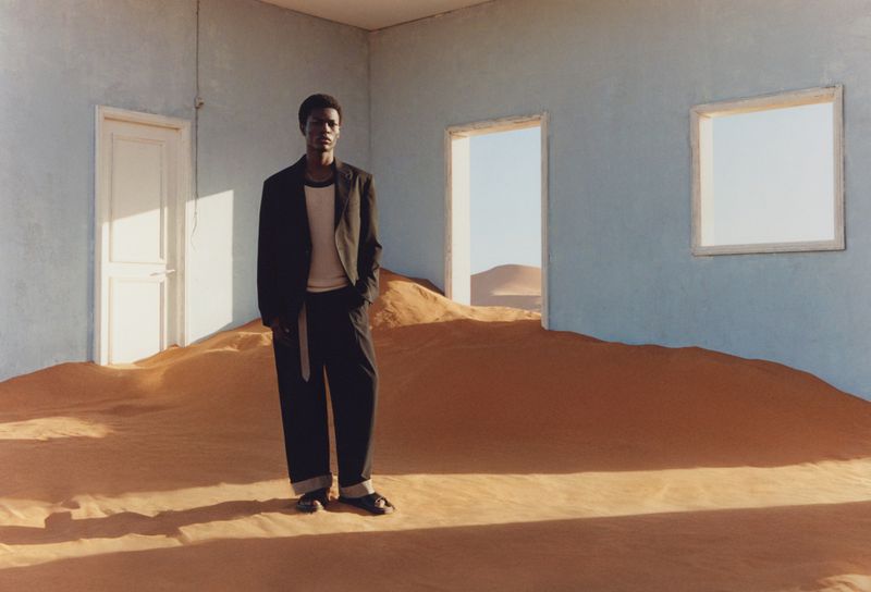 Black male model in a blazer stands on a desert dune inside an empty light blue room with two doors and a window.