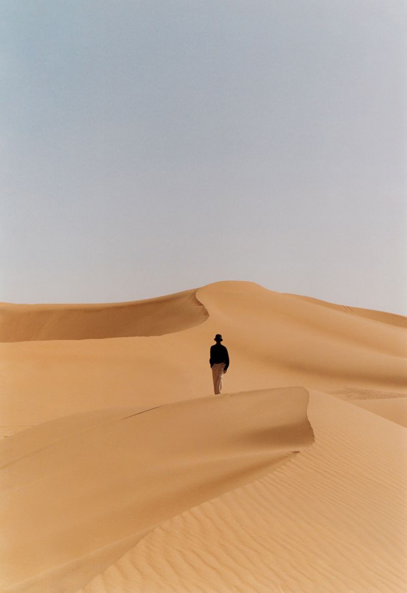 Person in hat walks alone on rippled sand dunes under a bright, overcast sky in a desert landscape setting during daytime.
