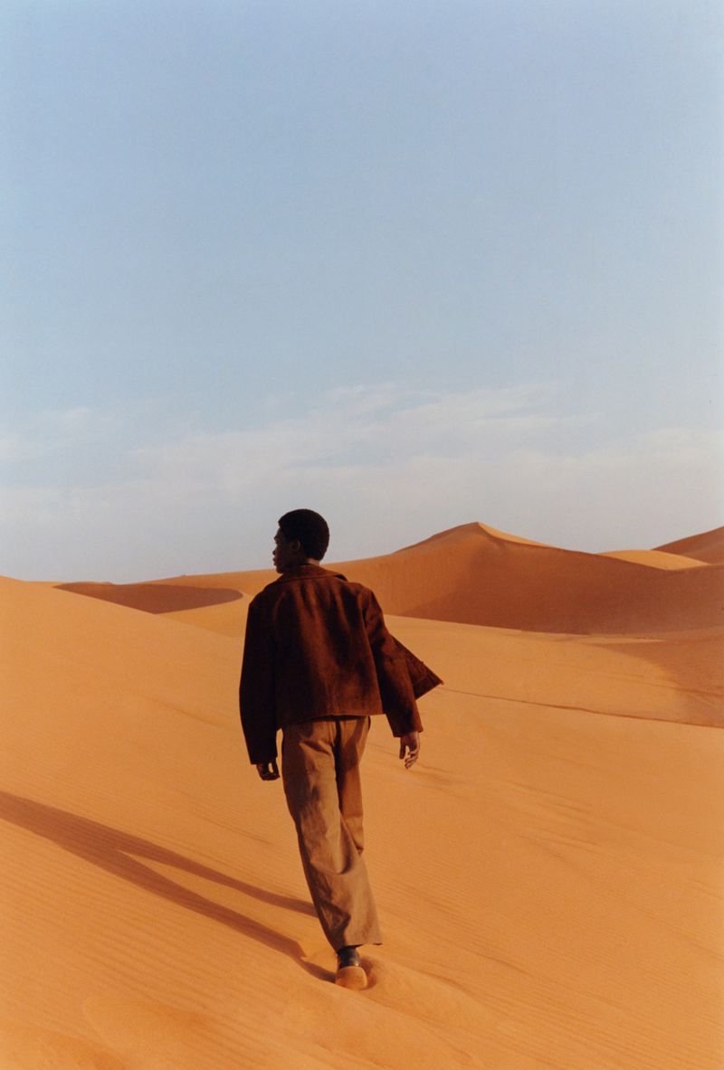 An African man walks through sand dunes wearing a jacket and pants, the blue sky is above. Landscape, natural light, and fashion.
