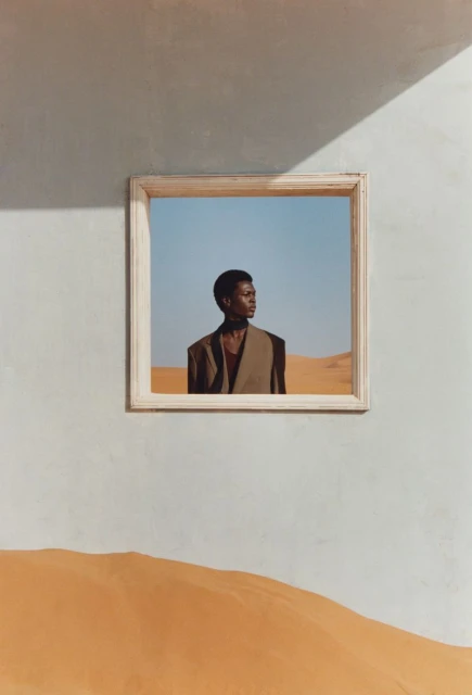 A striking fashion portrait features an African male model in a tan suit within a suspended frame, set against a surreal desert backdrop.
