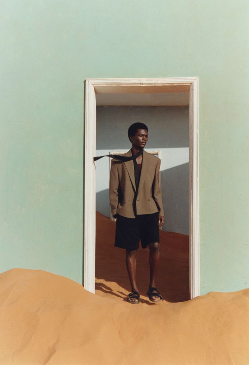 A Black male model in a beige blazer and black shorts stands in a doorway on a sand dune under a bright sky, looking off to the right.
