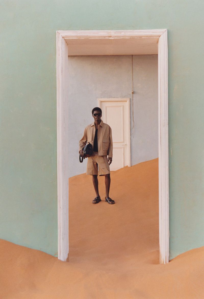 Full-length portrait of a male model standing in a doorway on sand wearing a beige jacket and shorts, photographed by Lukasz Pukowiec for Zara.