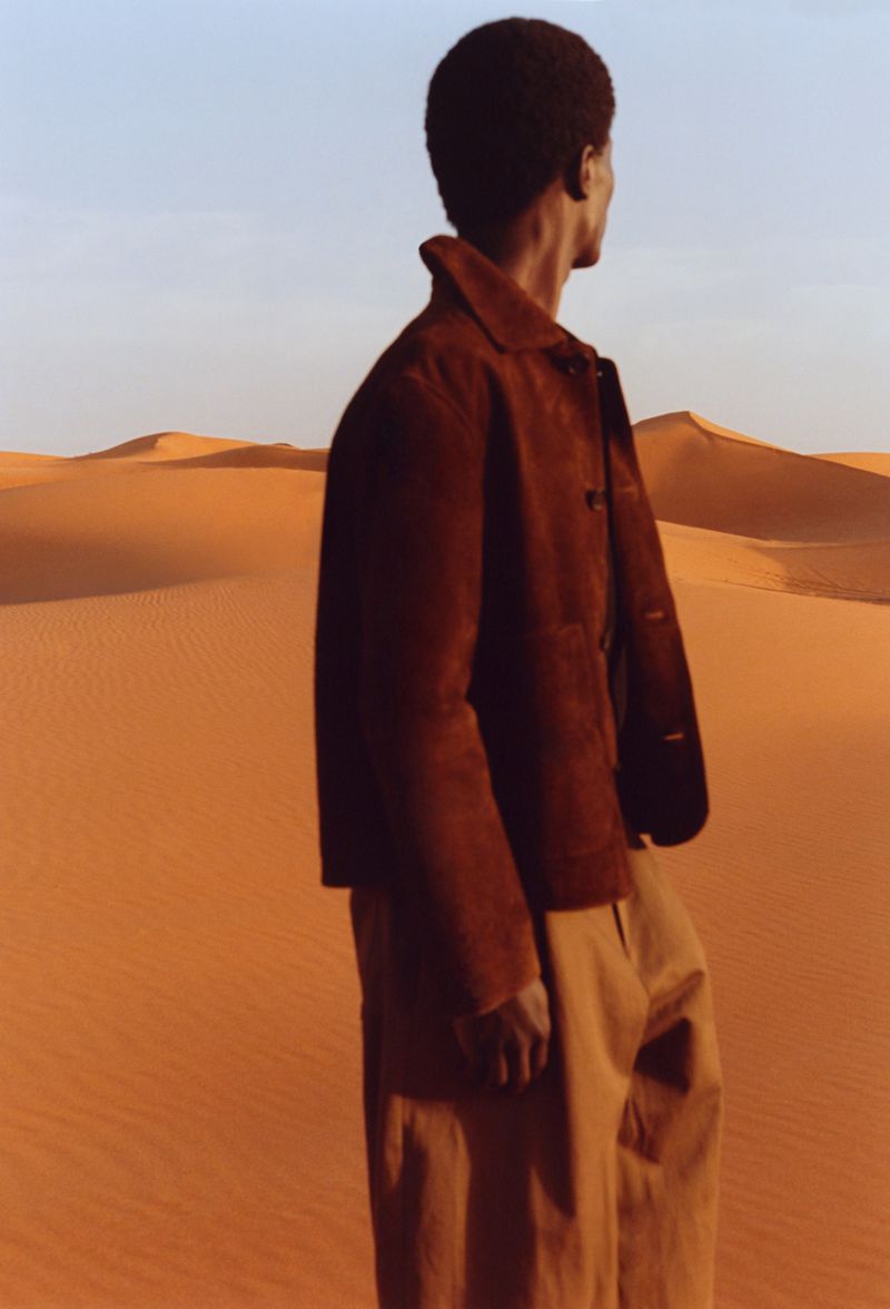 Model wearing a brown suede jacket standing in a vast desert landscape with sand dunes under a clear sky.