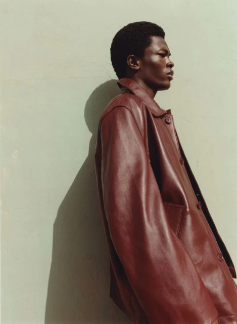 Portrait of a Black male model in profile, wearing a brown leather jacket against a pale green wall, casting shadow. Fashion editorial shot.