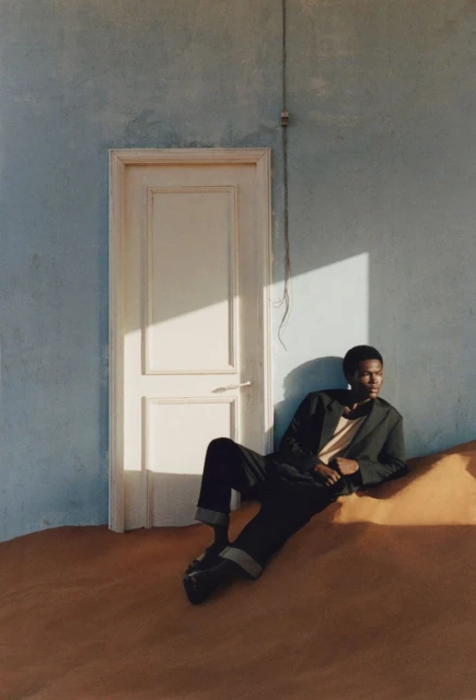 Adult Black man poses in a blazer and trousers in a minimalist setting with blue wall, white door, and sunlit brown floor.