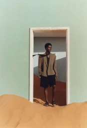 Model wearing a tan blazer and black shorts standing in an open doorway surrounded by sand dunes for Zara Man