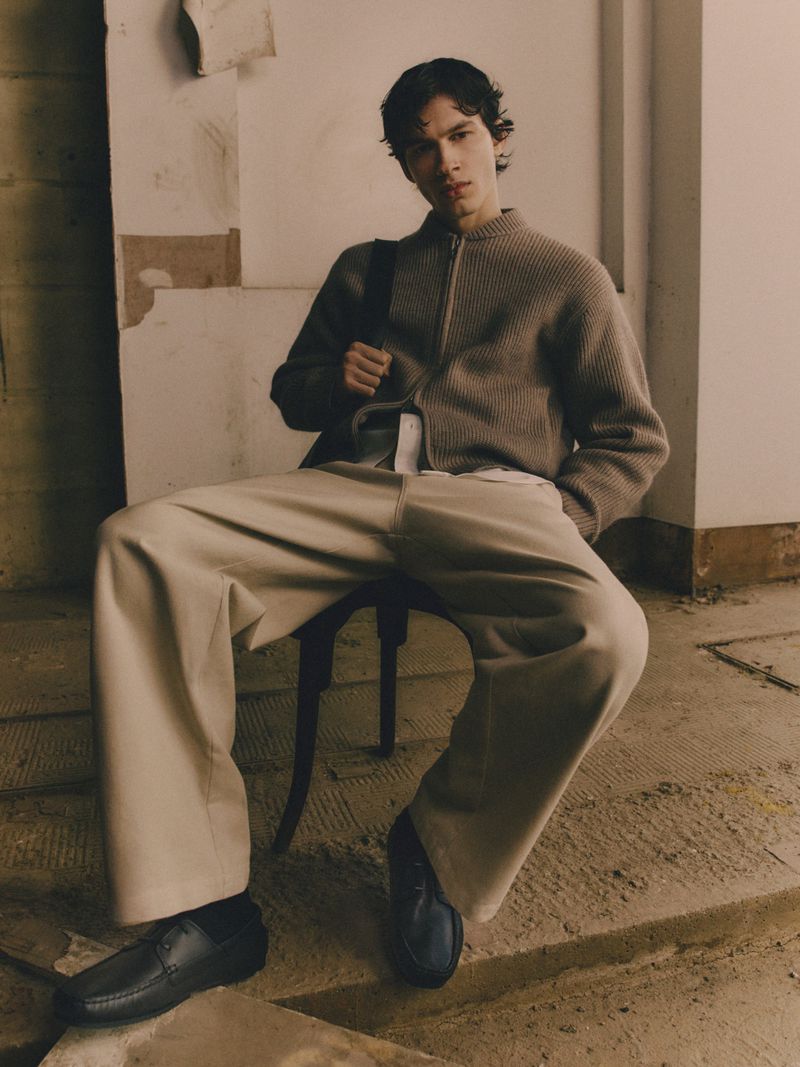 A stylish young man sits on a chair indoors wearing a cardigan, wide pants, and loafers, with a shoulder bag in a neutral color palette.