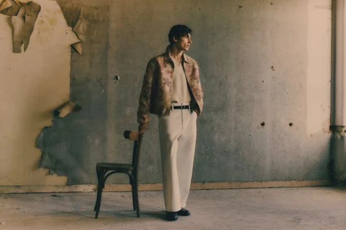 Adult male fashion model poses in beige suit jacket and pants in a sparse interior space, leaning on the back of a wooden chair.