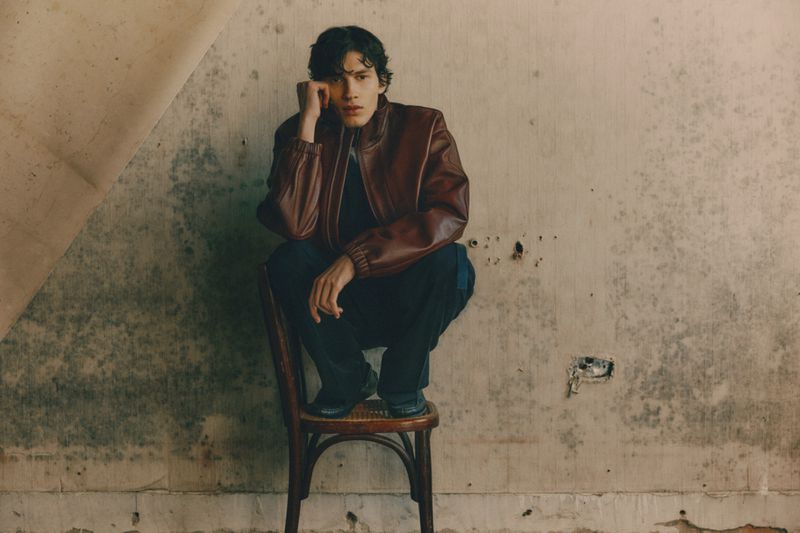 Model wearing a brown leather jacket and dark trousers crouched on a wooden chair against a weathered wall for Zara Tainted Neutrals
