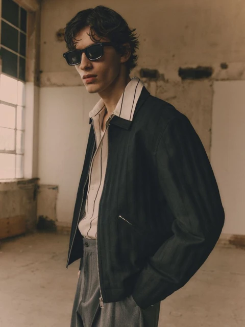 A young man with dark hair poses in sunglasses and a black jacket against a neutral indoor background, showcasing modern fashion.