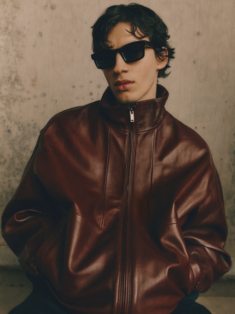 A dark-haired man with sunglasses poses in a studio, wearing a brown leather jacket with a zipper, against a neutral mottled backdrop.