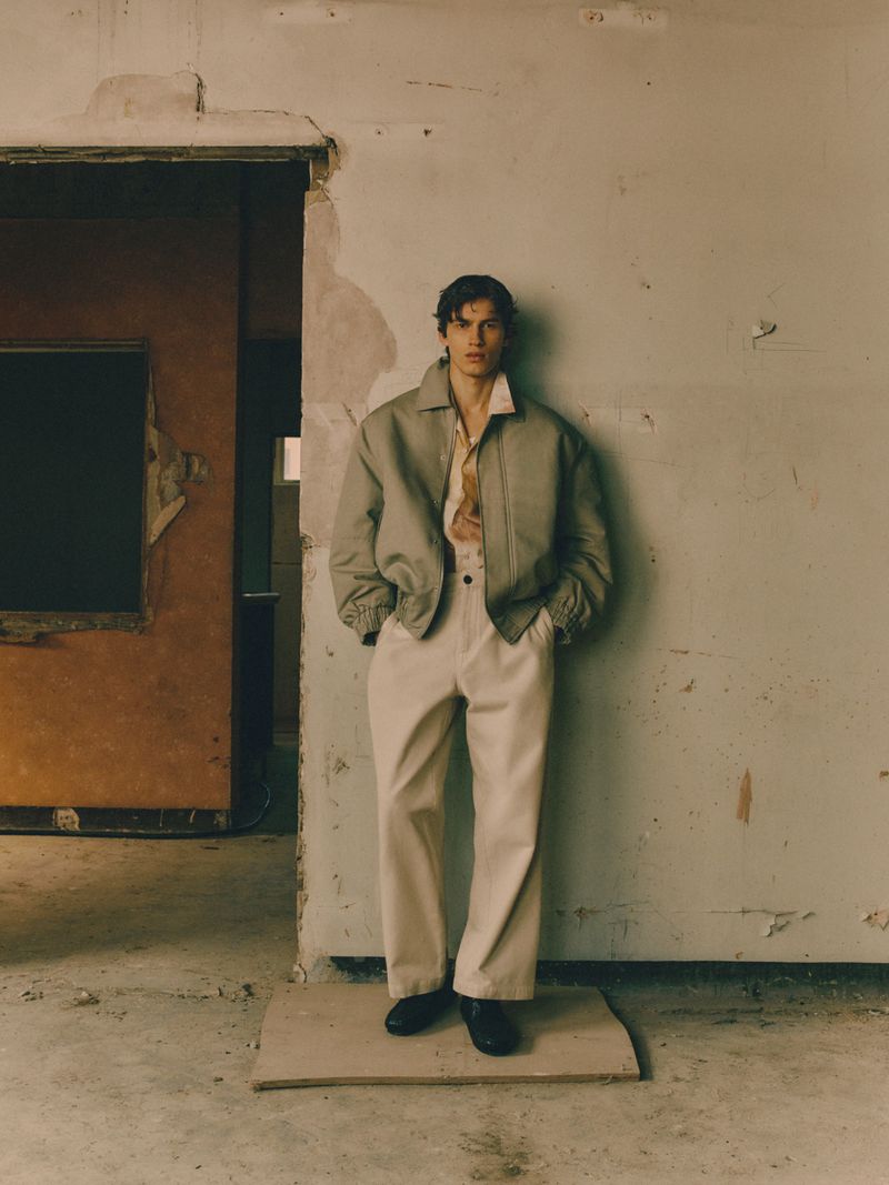 Man in jacket and trousers poses in weathered room. Fashion shot with neutral tones, casual style, and rough interior backdrop.