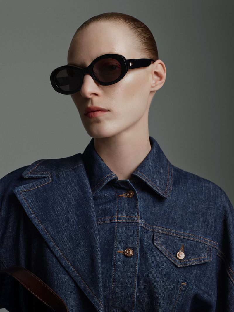 Close-up studio shot of a woman wearing a denim jacket and black sunglasses against a grey background in a fashion style.