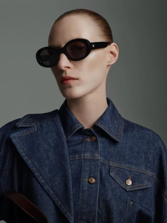 Close-up studio shot of a woman wearing a denim jacket and black sunglasses against a grey background in a fashion style.