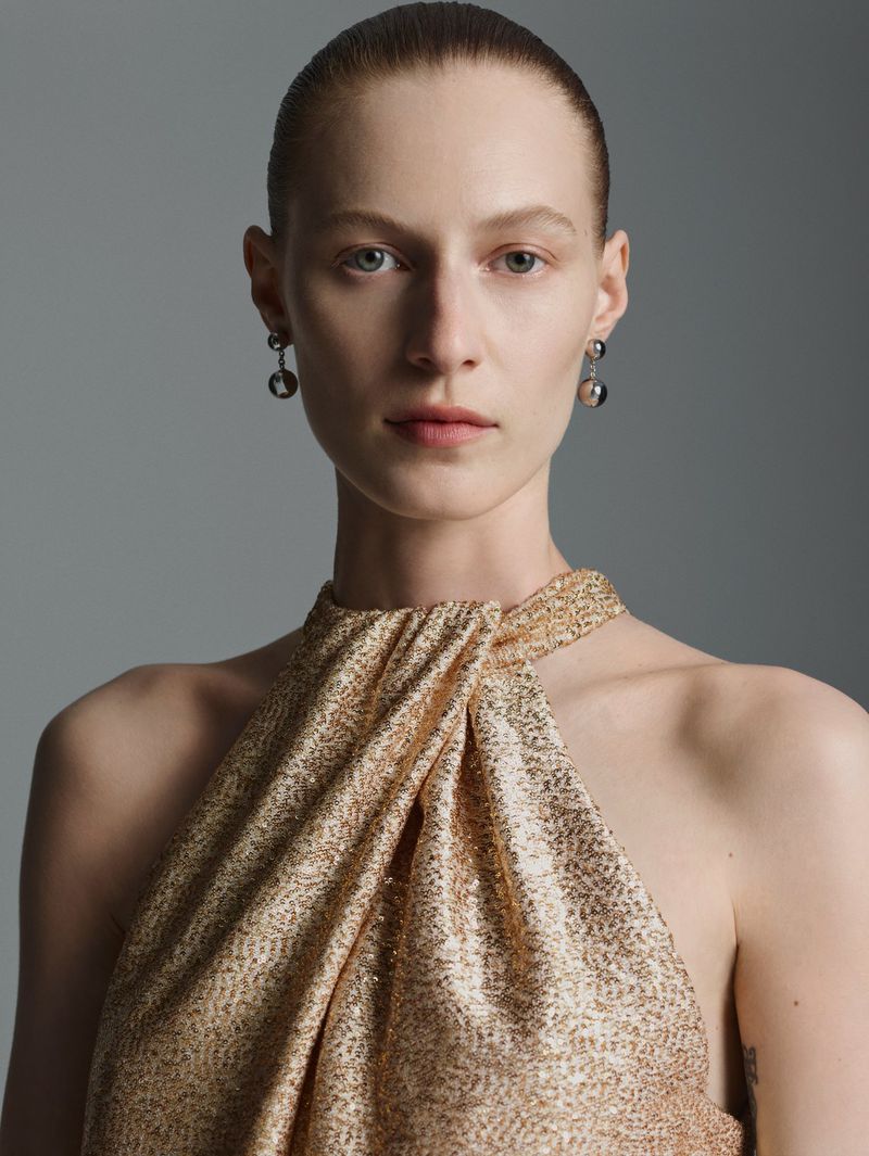 Close-up of a fair-skinned woman with hair slicked back, wearing a shimmering gold halter dress and grey earrings, against a grey backdrop.