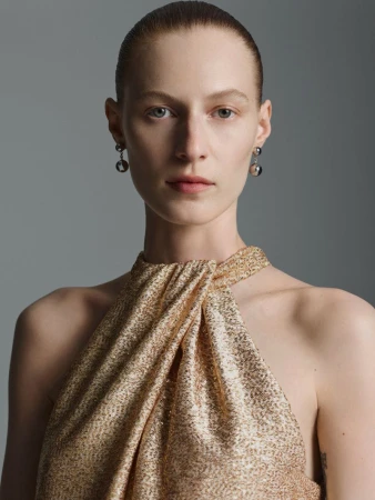 Close-up of a fair-skinned woman with hair slicked back, wearing a shimmering gold halter dress and grey earrings, against a grey backdrop.