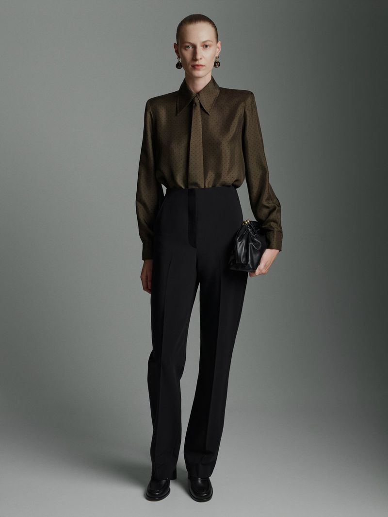 Full-length studio shot of a stylish adult woman wearing a brown silk blouse, black dress pants, and black heeled shoes with a black bag.