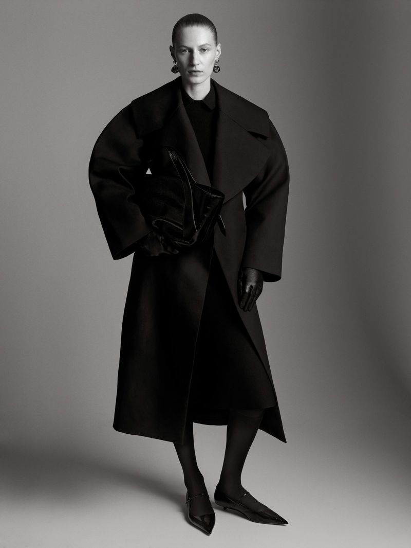 A model poses in a full-length shot wearing a dark coat, dress, tights, and shoes against a grey backdrop in a fashion studio setting.