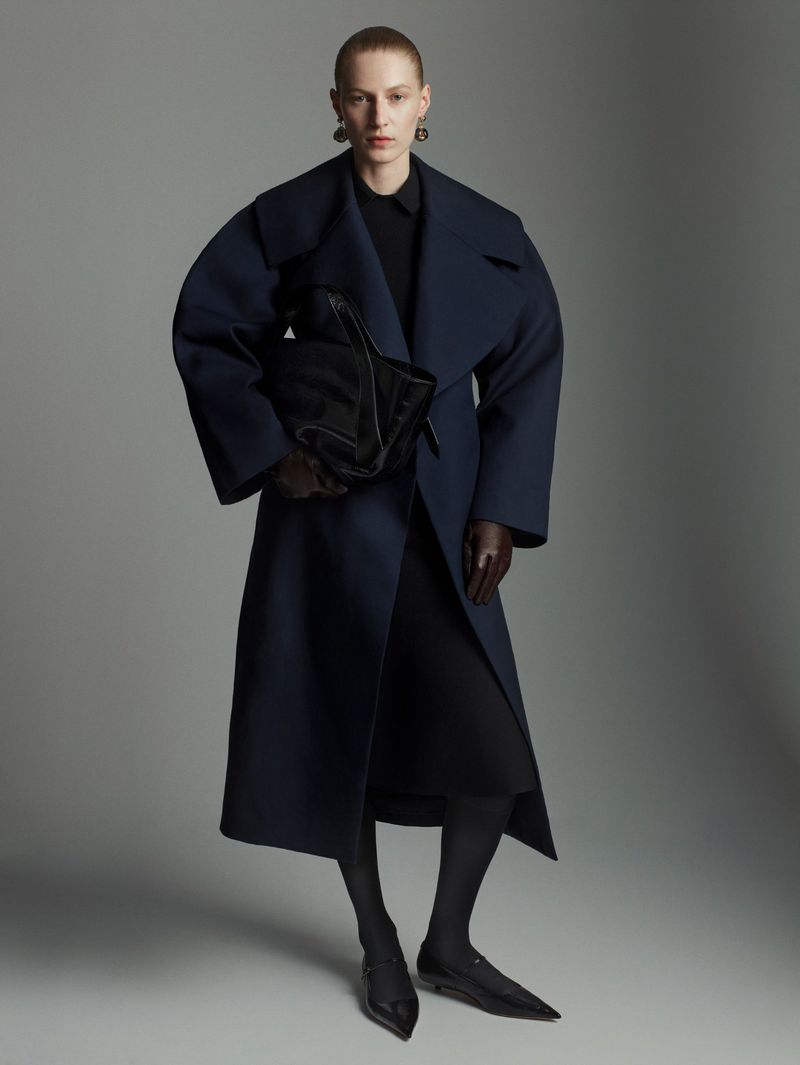 Fashion portrait of an adult female wearing a navy wrap coat with a black dress, tights, and leather flats in a studio.