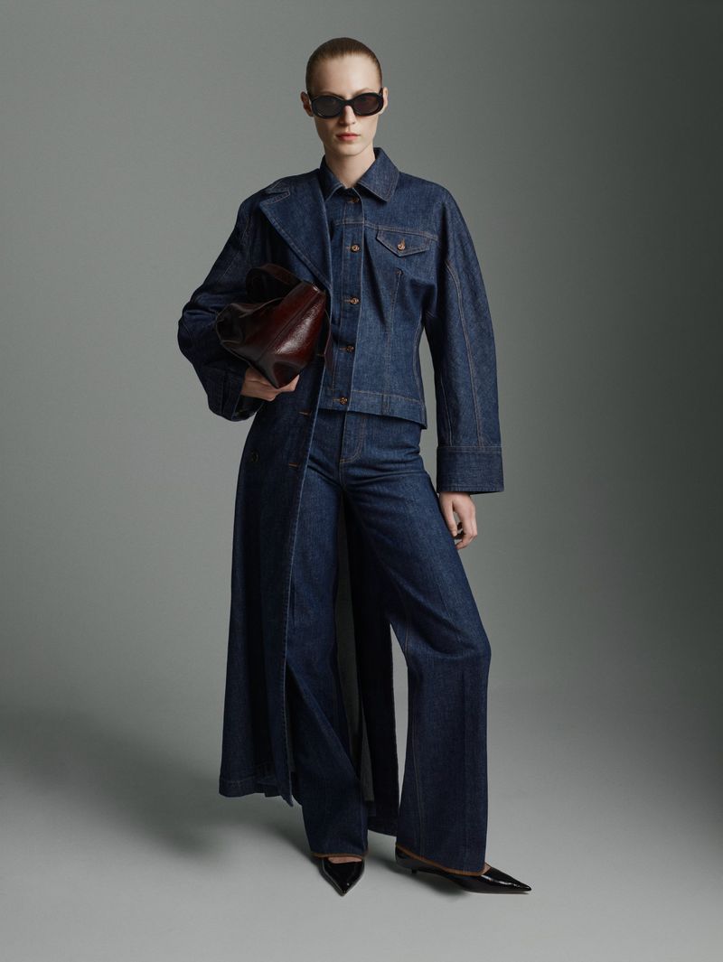 Woman in denim jacket, flared jeans, and long vest, holding a bag, wearing sunglasses, posing in a studio, fashion shot.