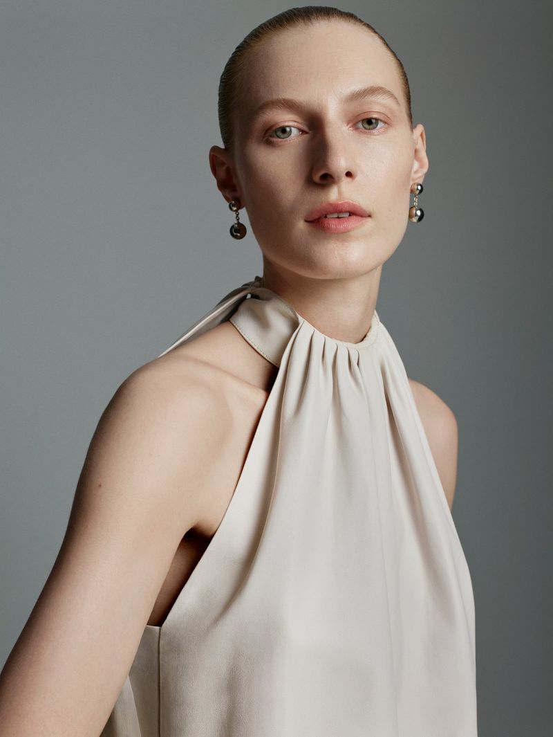 Close-up of a fair-skinned adult woman in a sleeveless top, showcasing her bare shoulders and elegant earrings against a gray backdrop.