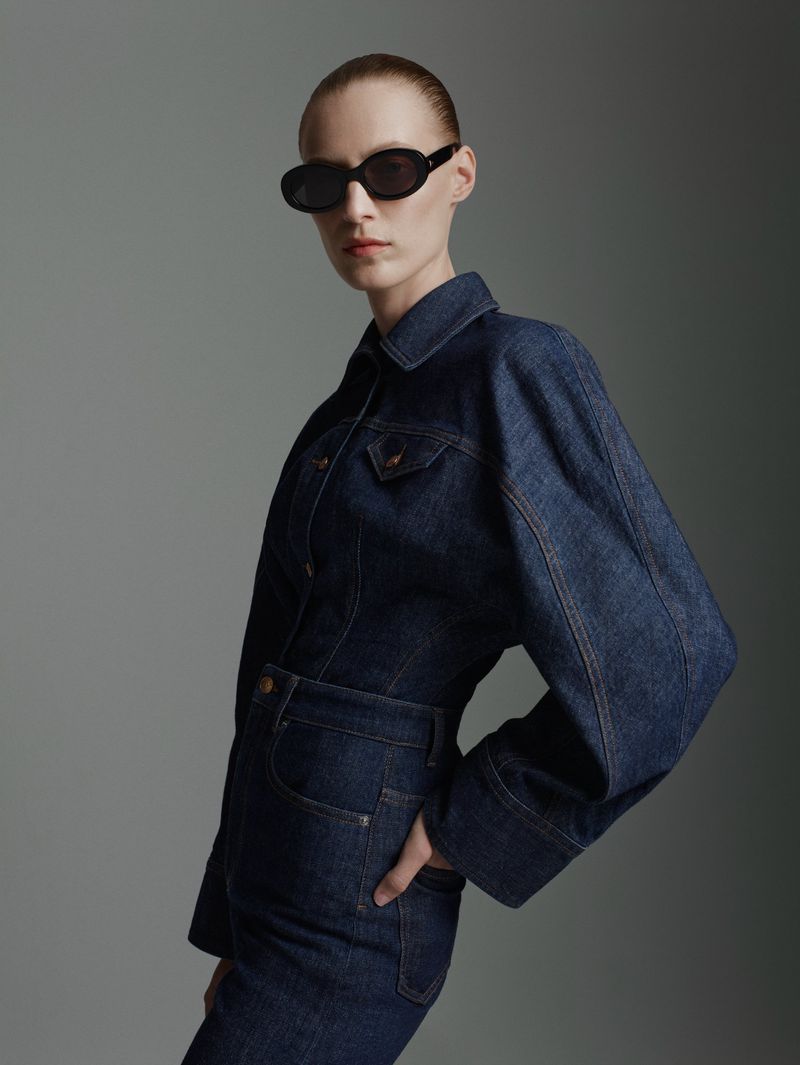 A fair-skinned woman in a denim jumpsuit and black oval sunglasses poses against a gray backdrop in a fashion-style studio portrait.