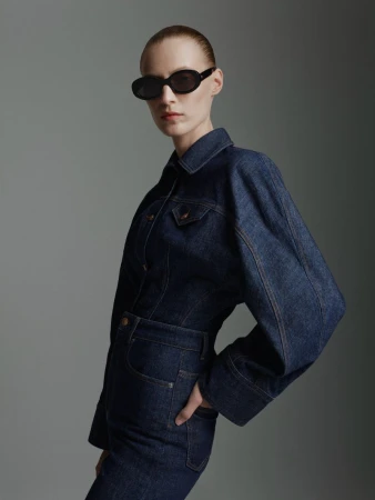 A fair-skinned woman in a denim jumpsuit and black oval sunglasses poses against a gray backdrop in a fashion-style studio portrait.