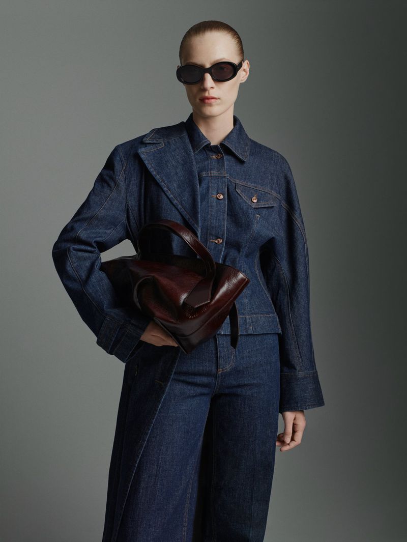 Fashionable adult woman in stylish denim jacket and wide-leg jeans, accessorized with sunglasses and a modern brown leather handbag.