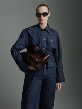 Fashionable adult woman in stylish denim jacket and wide-leg jeans, accessorized with sunglasses and a modern brown leather handbag.